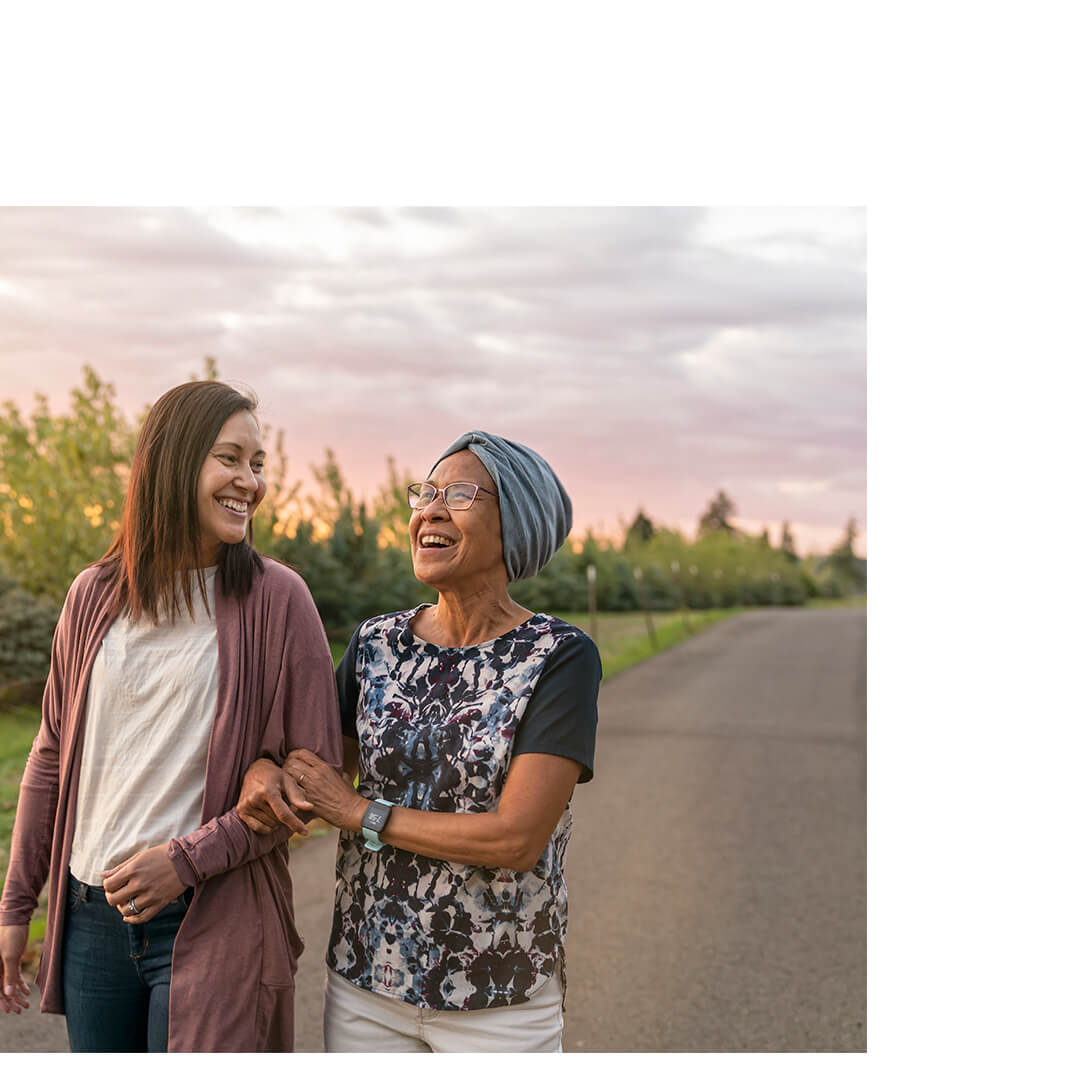 Two women, one a cancer survivor wearing a headscarf and her companion, walk outdoors at sunset, smiling and sharing a supportive moment