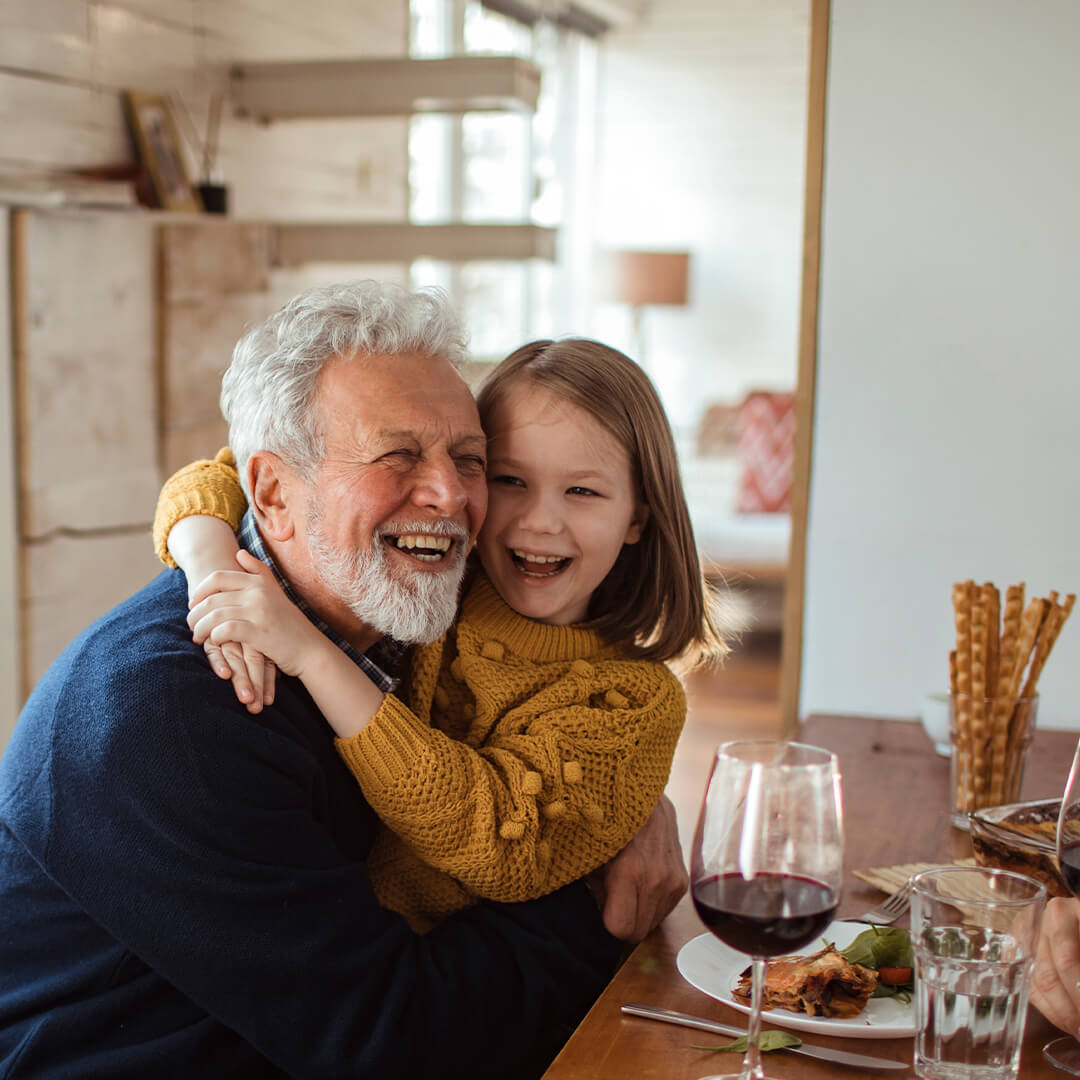 A young girl hugging her grandfather at the dinner table, both laughing and sharing a joyful moment