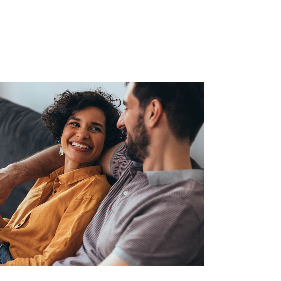 A smiling couple sits on a couch, discussing cancer risk prevention in a warm, sunny living-room setting