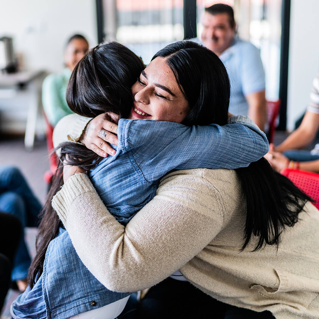 Two women embrace in a meeting room during a cancer support group session, conveying comfort and solidarity
